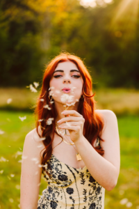 Young woman with long red hair in a patterned dress blowing dandelion seeds