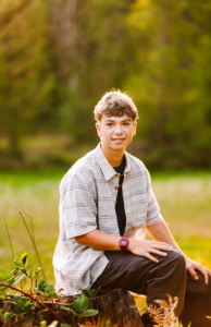 Teenage boy sitting on a tree stump in a striped shirt black pants and red wristwatch in a forest