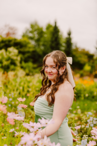 Young woman with wavy brown hair and a white bow in a light green strapless dress standing in wildflowers