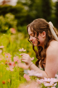 Young woman with wavy brown hair and a white bow kneeling in a garden touching pink flowers