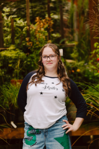 Young woman with long brown hair and glasses in a white and navy skater shirt and denim skirt hand on hip green plants behind her