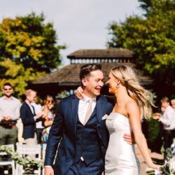 Bride and groom smiling at each other arm in arm as guests applaud at an outdoor ceremony
