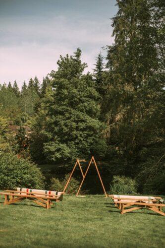A grassy outdoor area with wooden benches arranged on both sides facing a simple triangular wooden arch surrounded by lush green trees under a partly cloudy sky Snohomish Wedding Photography A grassy outdoor area with wooden benches arranged on both sides, facing a simple triangular wooden arch, surrounded by lush green trees under a partly cloudy sky. Snohomish Wedding Photography