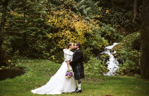 A bride in a white gown and groom in a kilt embrace outdoors in front of trees and a small waterfall with the bride holding a bouquet of purple flowers Snohomish Wedding Photography A bride in a white gown and groom in a kilt embrace outdoors in front of trees and a small waterfall, with the bride holding a bouquet of purple flowers. Snohomish Wedding Photography