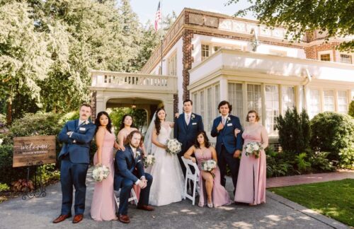 A wedding party poses outdoors in front of a large elegant house The bride stands in the center holding a bouquet surrounded by bridesmaids in pink dresses and groomsmen in blue suits Greenery and a welcome sign are visible Snohomish Wedding Photography A wedding party poses outdoors in front of a large, elegant house. The bride stands in the center holding a bouquet, surrounded by bridesmaids in pink dresses and groomsmen in blue suits. Greenery and a welcome sign are visible. Snohomish Wedding Photography