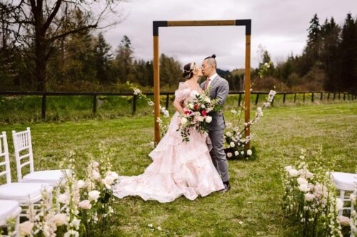 A couple embraces and kisses at an outdoor wedding ceremony standing in front of a wooden arch decorated with flowers The bride wears a pink floral gown and the groom is in a light gray suit White chairs and flowers line the aisle Snohomish Wedding Photography A couple embraces and kisses at an outdoor wedding ceremony, standing in front of a wooden arch decorated with flowers. The bride wears a pink floral gown, and the groom is in a light gray suit. White chairs and flowers line the aisle. Snohomish Wedding Photography