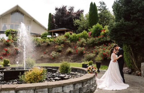 A bride and groom embrace and kiss beside a fountain in a landscaped garden with a bouquet on the ground and a large house in the background surrounded by greenery and colorful flowers Snohomish Wedding Photography A bride and groom embrace and kiss beside a fountain in a landscaped garden, with a bouquet on the ground and a large house in the background surrounded by greenery and colorful flowers. Snohomish Wedding Photography