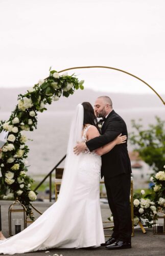 A bride and groom share a kiss under a circular floral arch at their outdoor wedding ceremony with water and greenery visible in the background Snohomish Wedding Photography A bride and groom share a kiss under a circular floral arch at their outdoor wedding ceremony, with water and greenery visible in the background. Snohomish Wedding Photography