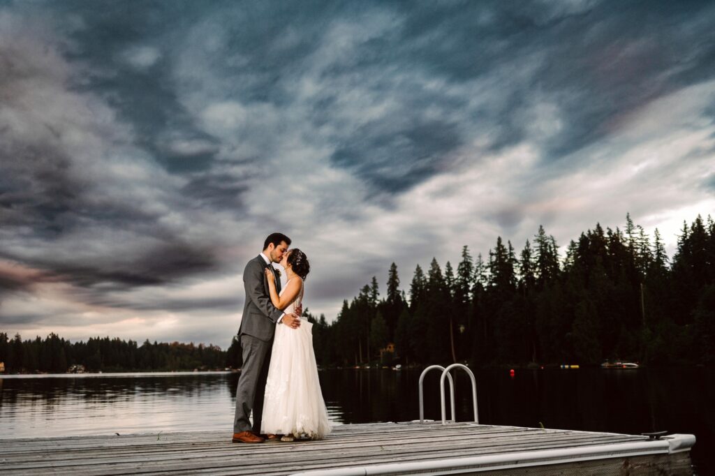 A bride and groom embrace on a wooden dock over a calm lake surrounded by tall trees and under a dramatic cloudy sky at dusk Snohomish Wedding Photography A bride and groom embrace on a wooden dock over a calm lake, surrounded by tall trees and under a dramatic, cloudy sky at dusk. Snohomish Wedding Photography