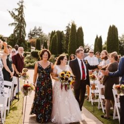 Bride holding a sunflower bouquet walking down an outdoor aisle with two people guests standing and smiling on both sides among trees