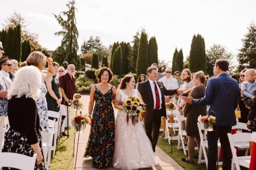 Lord Hill Farms A bride in a white dress holding a sunflower bouquet walks down an outdoor aisle with two people Guests stand and smile on either side surrounded by greenery and trees on a sunny day Snohomish Wedding Photography Lord Hill Farms A bride in a white dress holding a sunflower bouquet walks down an outdoor aisle with two people Guests stand and smile on either side surrounded by greenery and trees on a sunny day Snohomish Wedding Photography