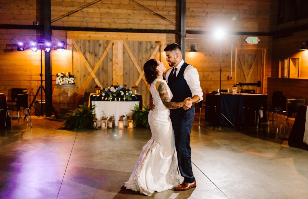 A bride and groom share a first dance in a warmly lit barn venue, smiling at each other. The bride wears a white dress and the groom a vest and tie. Decor includes plants, lanterns, and a “Mrs” sign in the background. Snohomish Wedding Photography