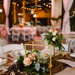A close up of a wedding table centerpiece with peach and white roses greenery and a gold geometric frame set on a draped beige tablecloth Blurred tables and string lights are visible in the warmly lit background Snohomish wedding photography