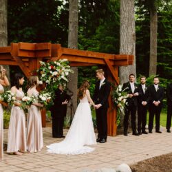 A bride and groom stand under a wooden arbor during an outdoor wedding ceremony holding hands Bridesmaids in champagne dresses and groomsmen in black suits stand on either side surrounded by tall trees and greenery Snohomish wedding photography