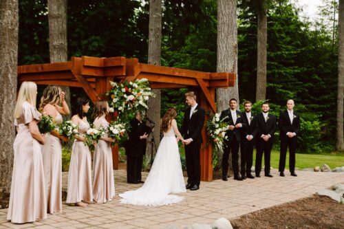 A bride and groom stand under a wooden arbor during an outdoor wedding ceremony holding hands Bridesmaids in champagne dresses and groomsmen in black suits stand on either side surrounded by tall trees and greenery Snohomish Wedding Photography A bride and groom stand under a wooden arbor during an outdoor wedding ceremony, holding hands. Bridesmaids in champagne dresses and groomsmen in black suits stand on either side, surrounded by tall trees and greenery. Snohomish Wedding Photography