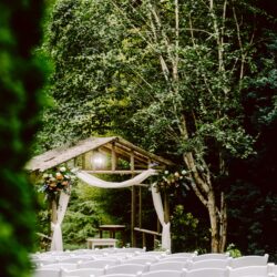 Rows of white chairs facing a wooden arbor decorated with flowers and white drapery set outdoors among green trees