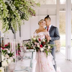 Bride in a floral pink gown and groom in a gray suit embracing beside a flower decorated reception table