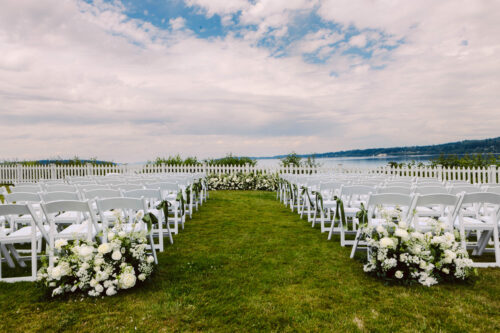Rows of white chairs arranged on grass face a floral wedding altar with white flowers along the aisle and a scenic view of water and mountains beyond a white picket fence under a partly cloudy sky Snohomish Wedding Photography Rows of white chairs arranged on grass face a floral wedding altar, with white flowers along the aisle and a scenic view of water and mountains beyond a white picket fence under a partly cloudy sky. Snohomish Wedding Photography