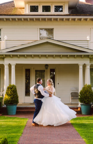 A bride and groom dance joyfully together outside a large white house with columns surrounded by green lawns and potted shrubs in warm sunlight Snohomish Wedding Photography A bride and groom dance joyfully together outside a large white house with columns, surrounded by green lawns and potted shrubs, in warm sunlight. Snohomish Wedding Photography