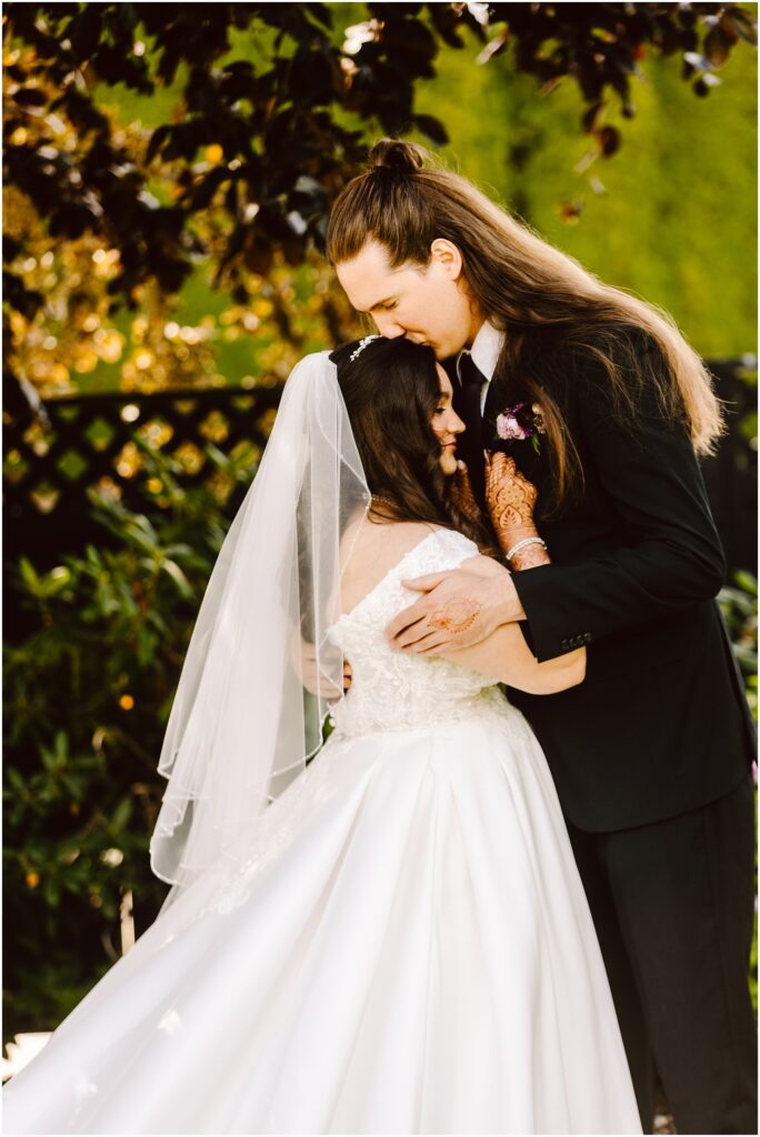 Bride and groom share a kissembrace outdoors bride in a lace wedding dress with veil and henna on her hands groom in a dark suit with boutonniere green background