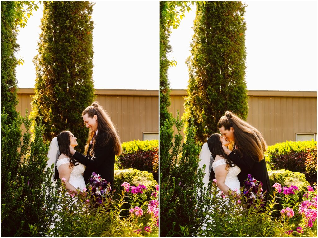 Bride in a lace wedding dress with veil and groom in a dark suit share a kiss in a sunlit garden with pink flowers and tall shrubs framing them