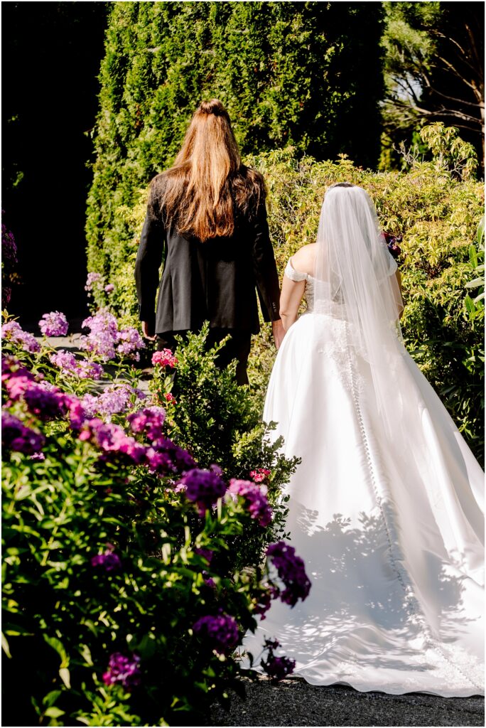 Bride in a white wedding gown and veil walks beside a man in a black suit along a garden path with vibrant purple flowers