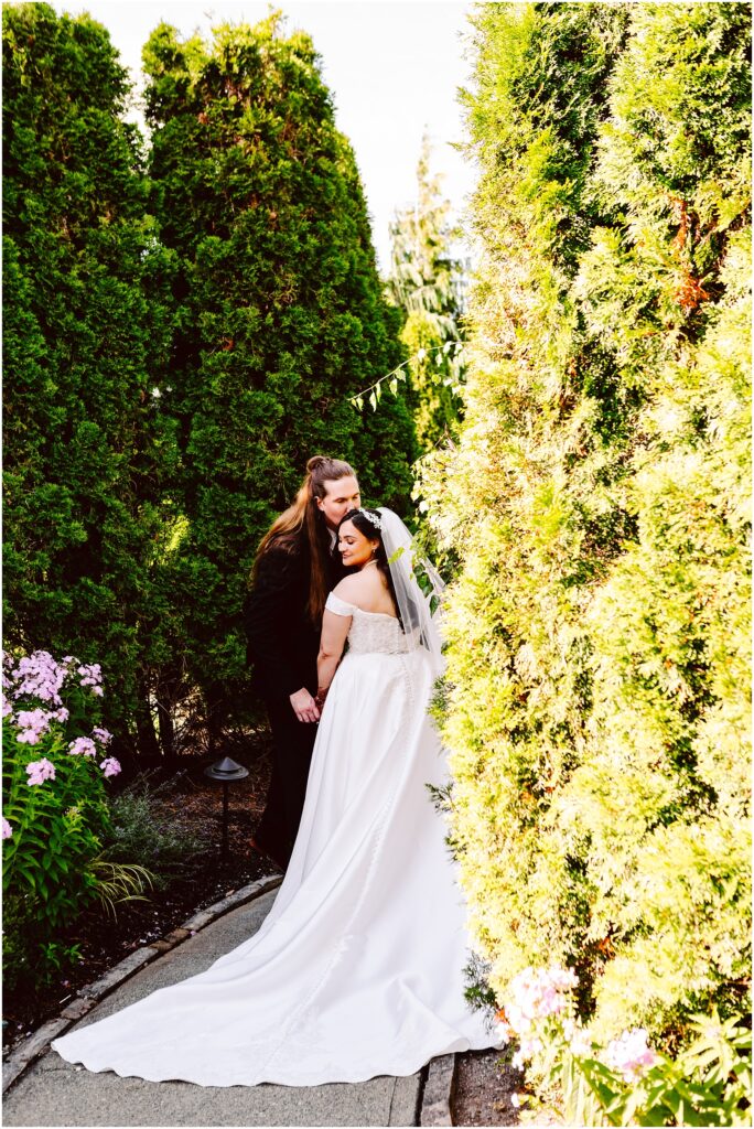 Bride in a white wedding gown and groom in a black suit kiss in a garden framed by tall green hedges and flowering plants on a sunny day