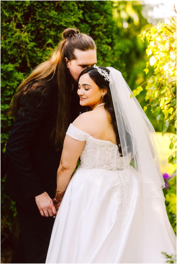 Bride in a lace wedding dress and veil stands with a long haired groom in a garden as he kisses her head and they hold hands