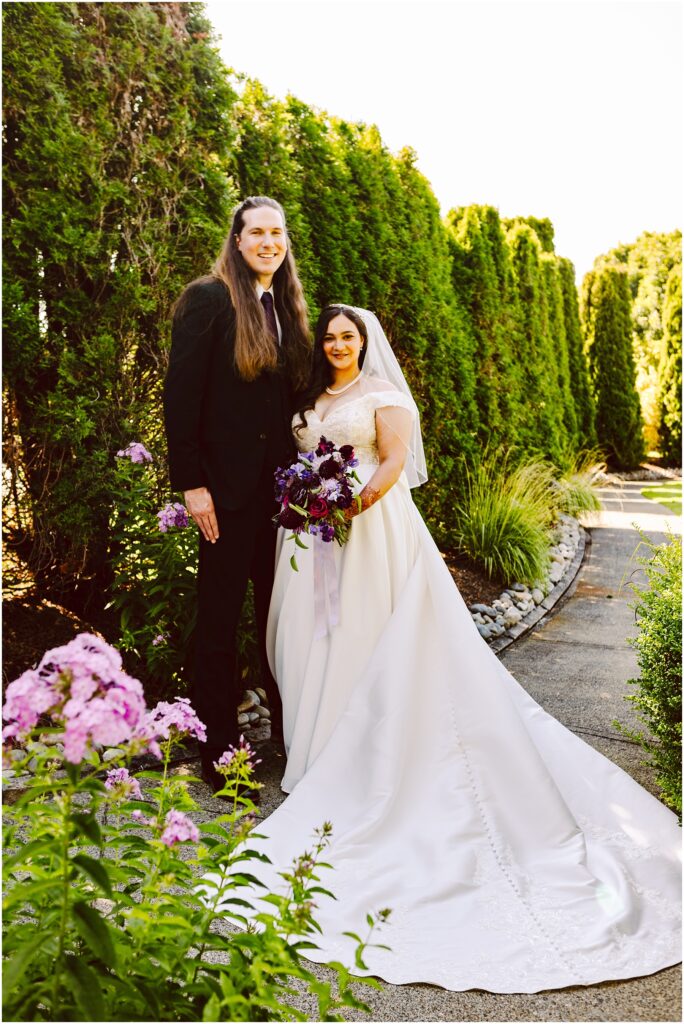 Bride in a white wedding gown and veil holding a purple bouquet stands next to a man in a black suit outdoors along a hedge lined path smiling together