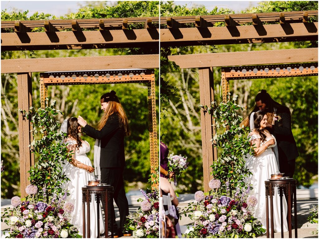 Bride in a white gown with veil stands under a wooden arbor as a man in a black suit adjusts her hair with lush flower arrangements nearby