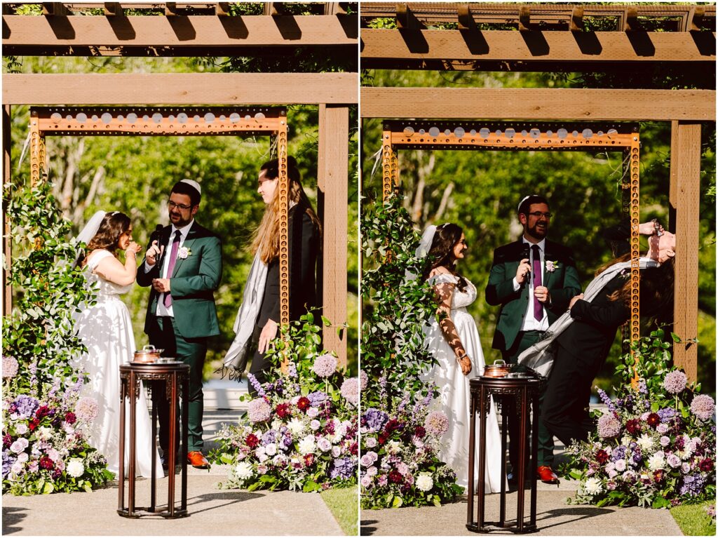 Bride in a white gown and groom in a green suit stand under a wooden arbor the groom speaking into a microphone as a woman in black and veil looks on colorful flowers frame the ceremony in the foreground