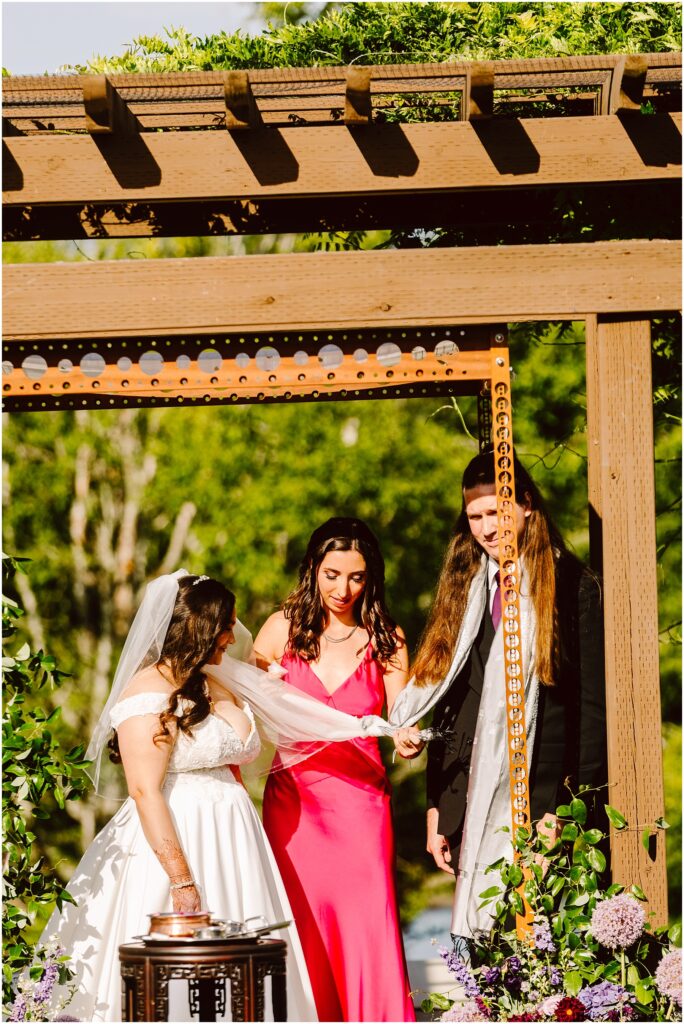 Bride in a white wedding dress with veil standing under a wooden arbor while a woman in a pink dress helps adjust her veil outdoors with flowers nearby