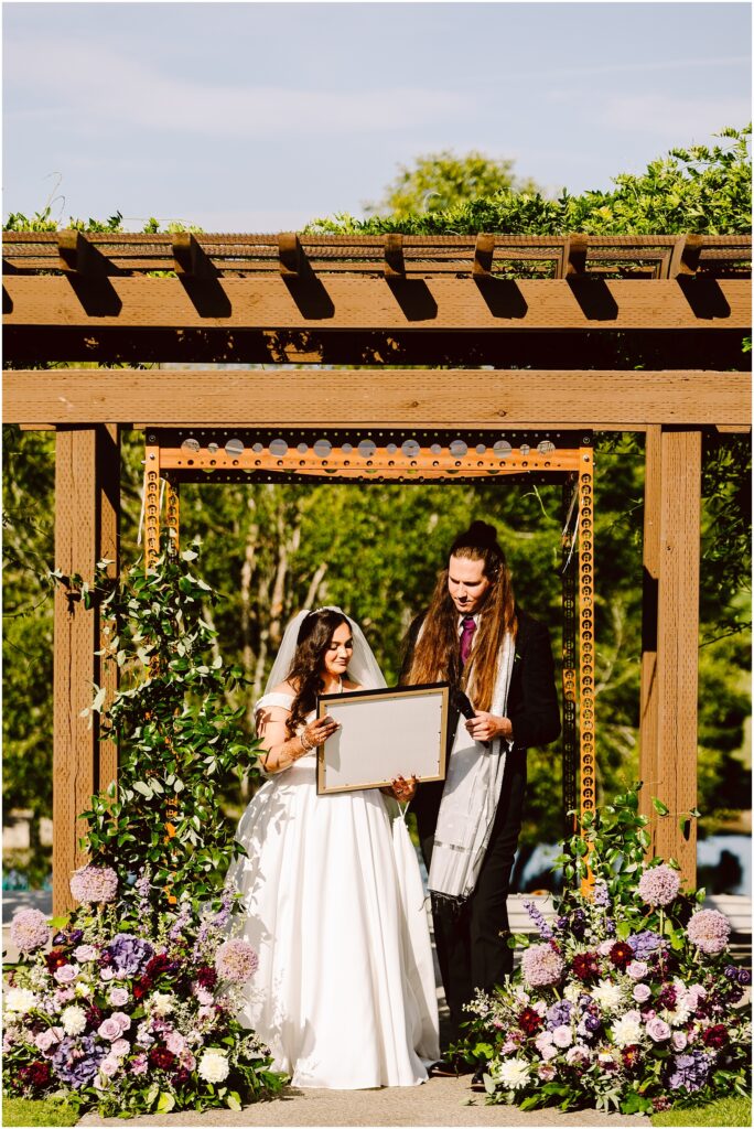Bride and groom under a wooden pergola the bride holding a framed board amid vibrant pink purple and white flowers