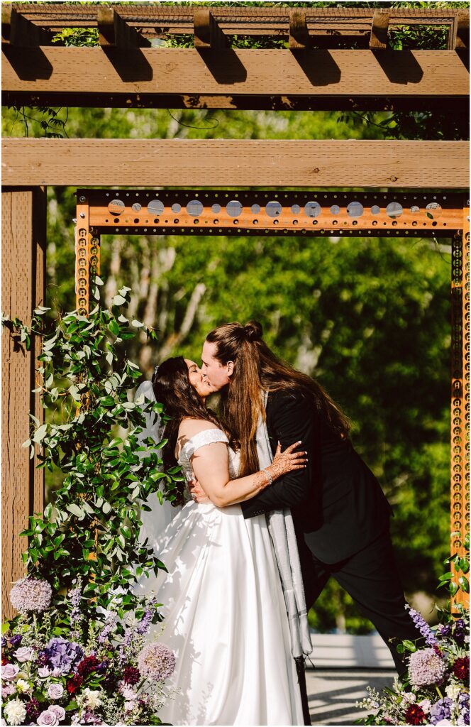 Two brides kiss beneath a rustic wooden arch framed by greenery and purple flowers at an outdoor wedding start scene