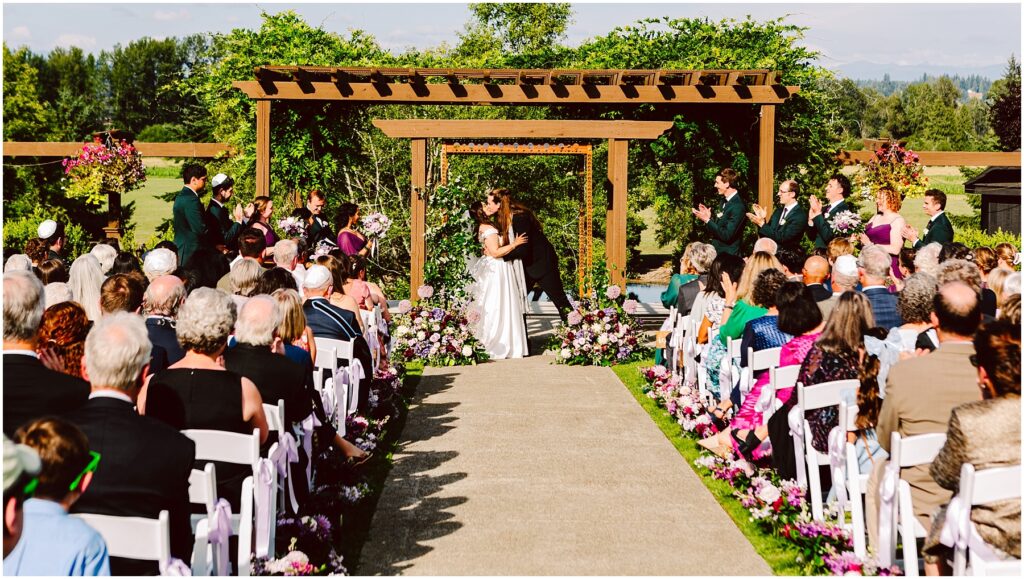 Bride and groom kiss under a wooden arbor at an outdoor wedding surrounded by rows of seated guests and floral arrangements on the aisle sides