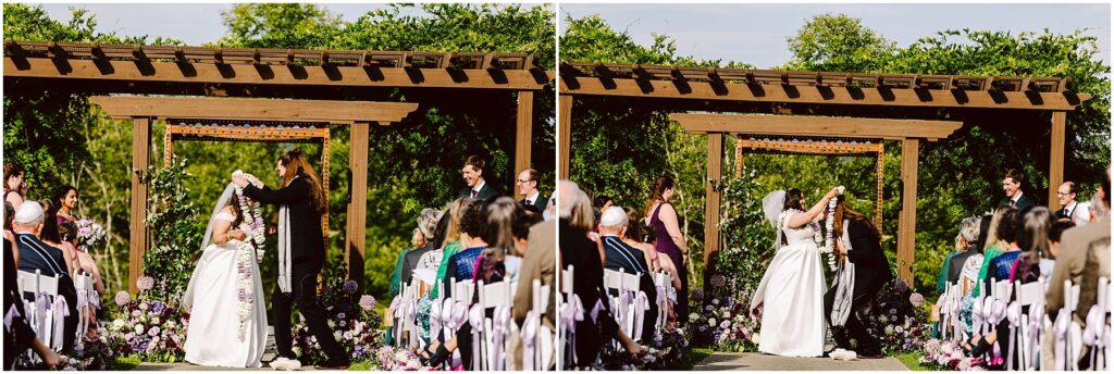 Outdoor wedding ceremony under a wooden arbor with a bride in a white dress and guests seated along the aisle flowers lining the aisle and decorations in purple and white