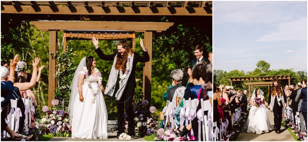 Bride in a white gown and groom in a dark suit wearing flower garlands walk under a wooden arch as guests cheer and clap Flowers line the aisle on both sides