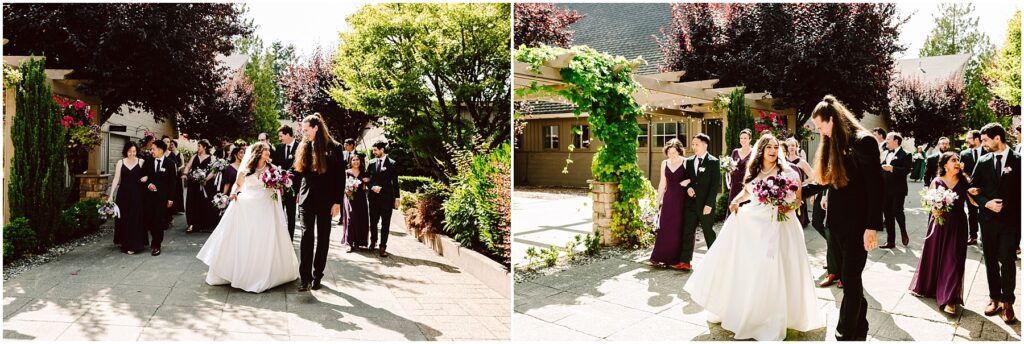 Bride in a white gown with a pink bouquet walks with the groom as bridesmaids in burgundy and groomsmen in black follow along a sunlit garden path
