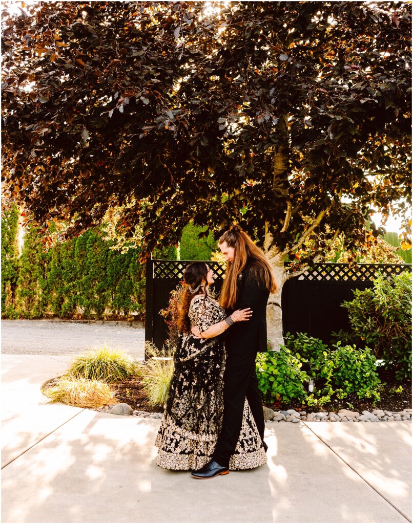Woman in a black formal dress hugs a girl in a gold embroidered gown outdoors under a large tree