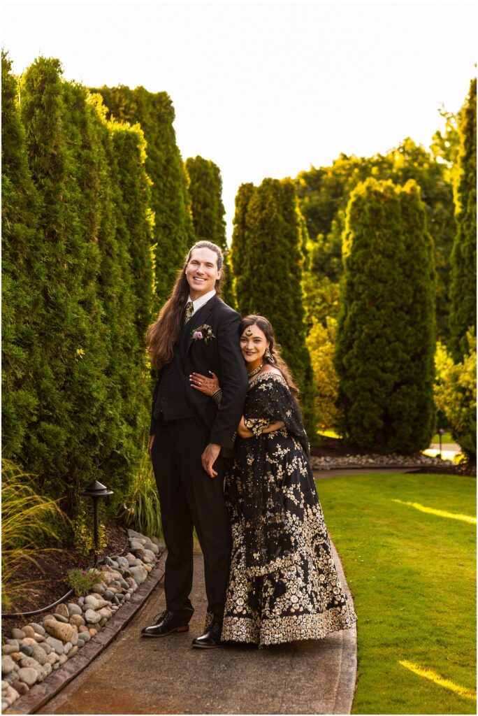 Smiling couple in formal attire standing arm in arm on a garden path with tall green hedges on both sides