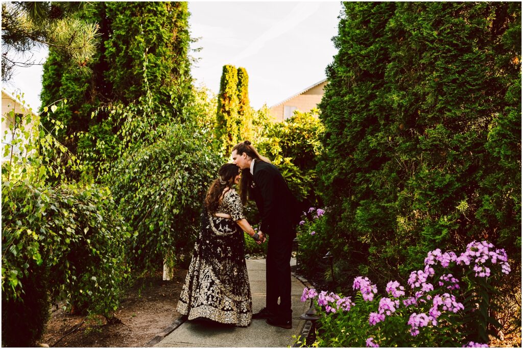 A couple dressed in formal attire holds hands in a sunlit garden path surrounded by lush green hedges and pink flowers