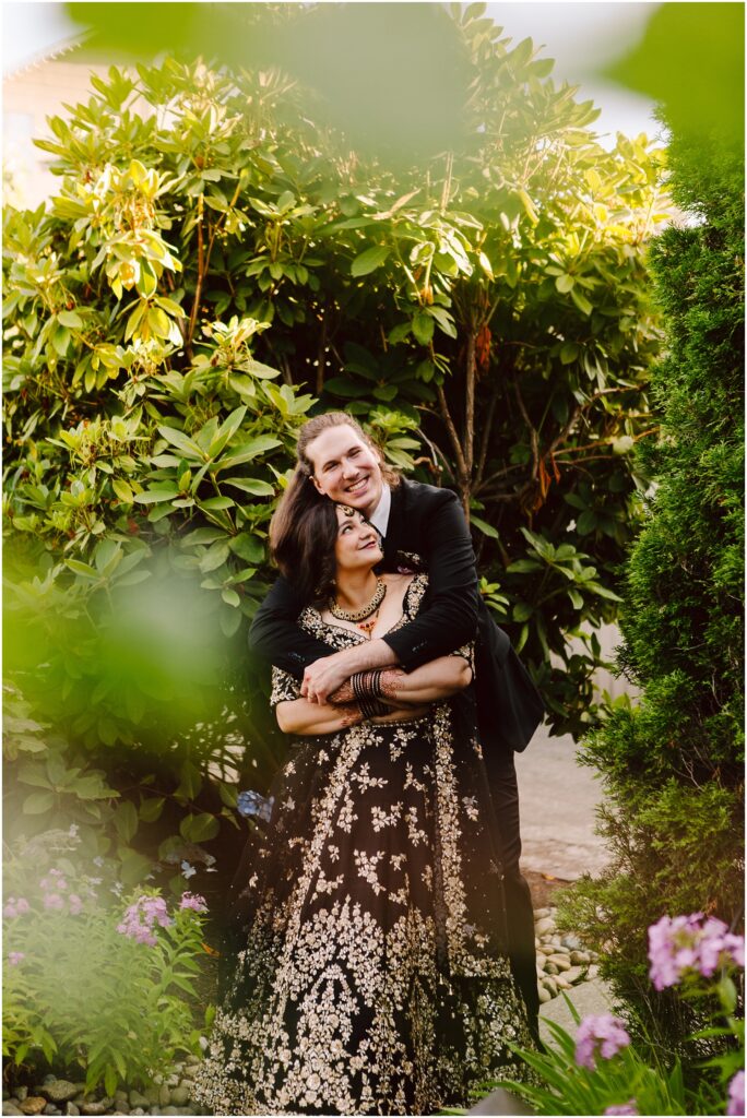 A couple poses arm in arm in a garden among dense green shrubs the woman in a black embroidered dress and the man in a suit smiling at the camera