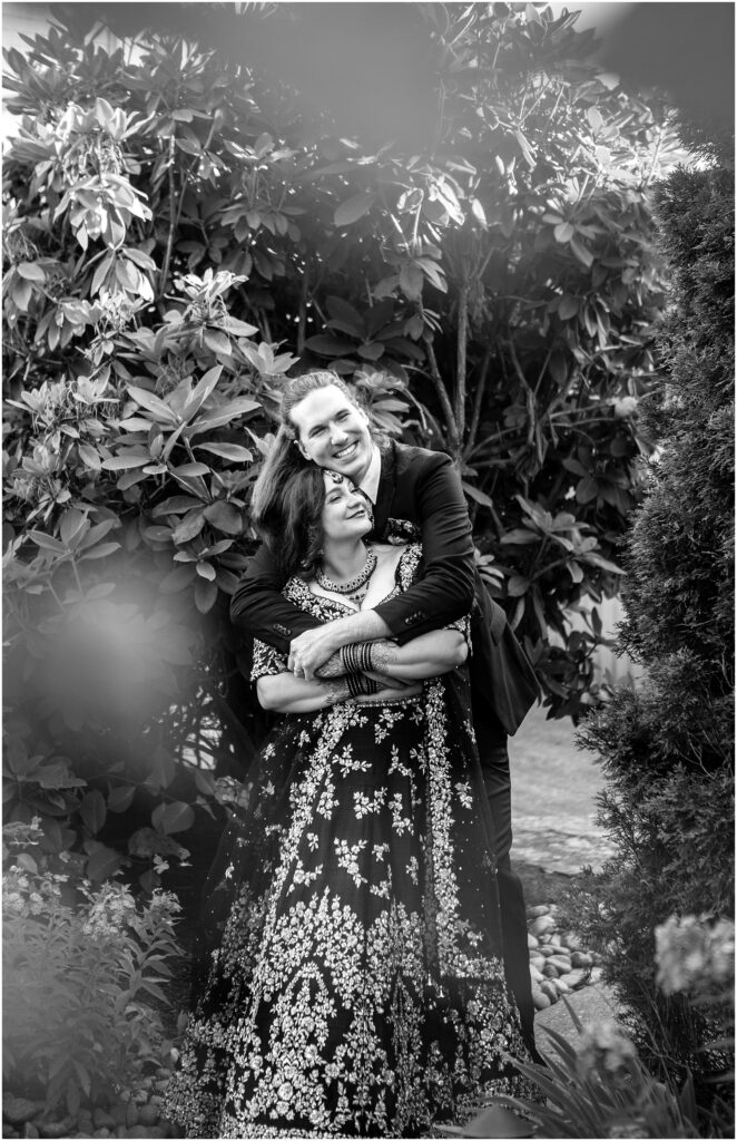 Smiling couple embracing in a garden man in a suit behind a woman in a heavily embroidered dress both looking happy black and white