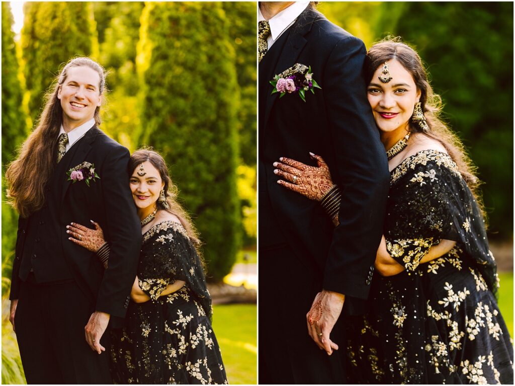 Smiling couple outdoors a man in a black suit with boutonniere and a woman in a gold embroidered black outfit she rests her head on his arm with henna on her hands