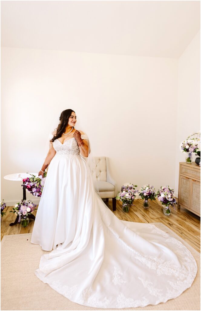Bride in a white wedding gown with a long train and veil standing in a bright room with flowers around her
