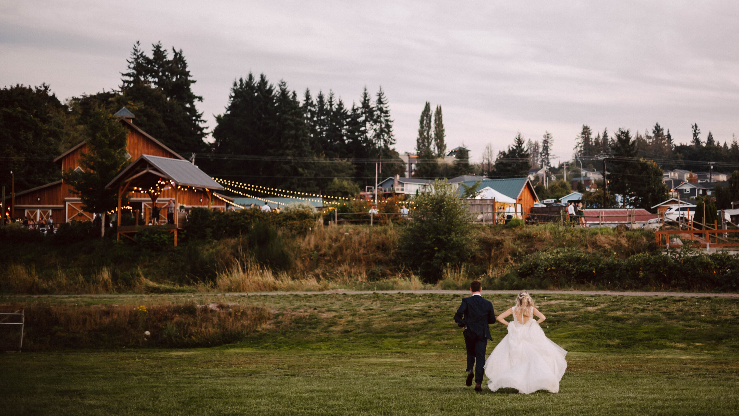 Bride and groom run across a grassy field toward a rustic barn wedding venue lit by string lights at dusk