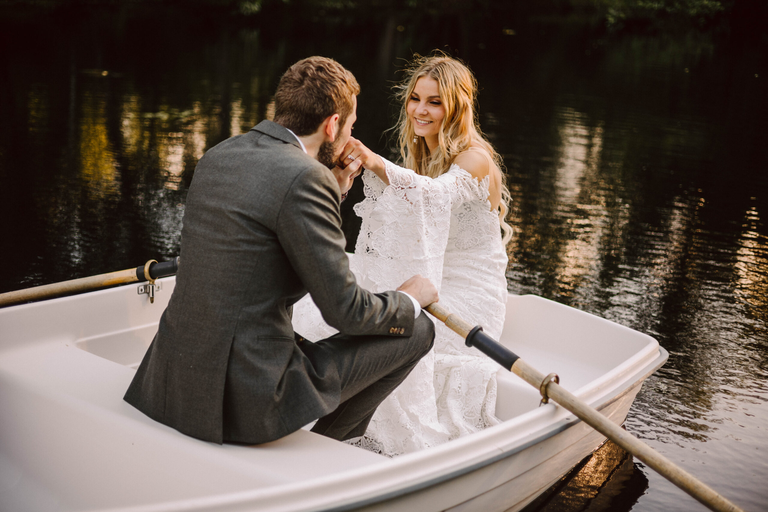 A man in a gray suit rows a small white boat as a woman in a white lace dress smiles at him on a calm lake