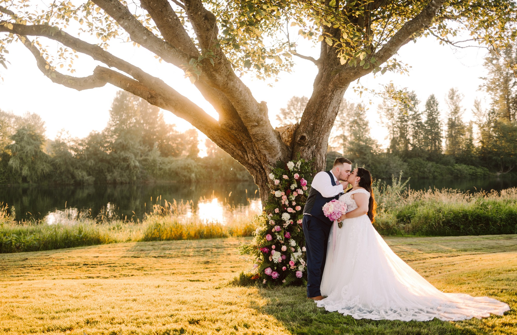 Bride in a white gown and groom in a dark suit kiss beside a large floral tree arch by a calm lakeside at sunset