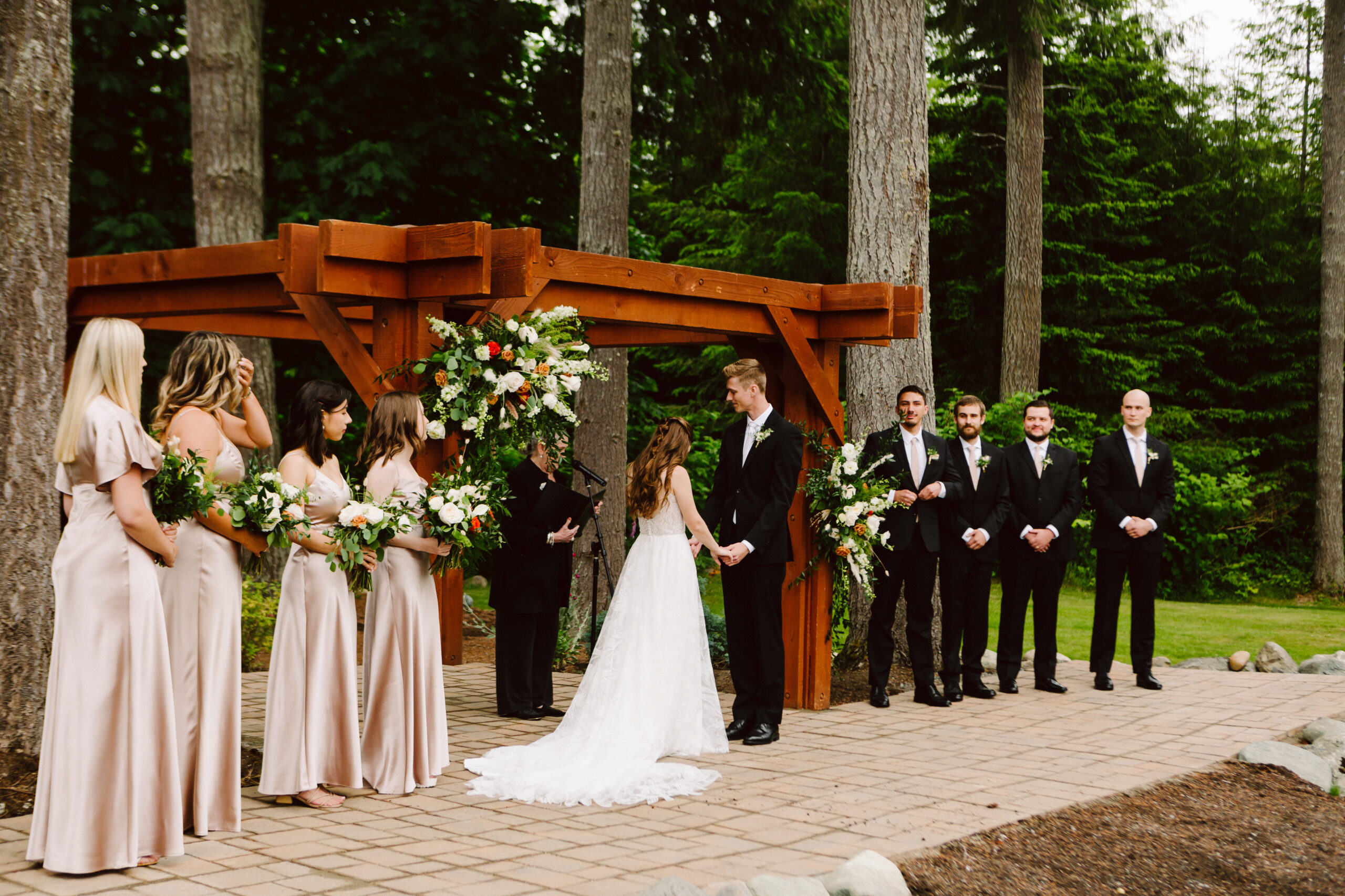 Bride and wedding party under a wooden arch with floral arrangements bridesmaids in blush dresses on the left groomsmen in black suits on the right forest backdrop behind them