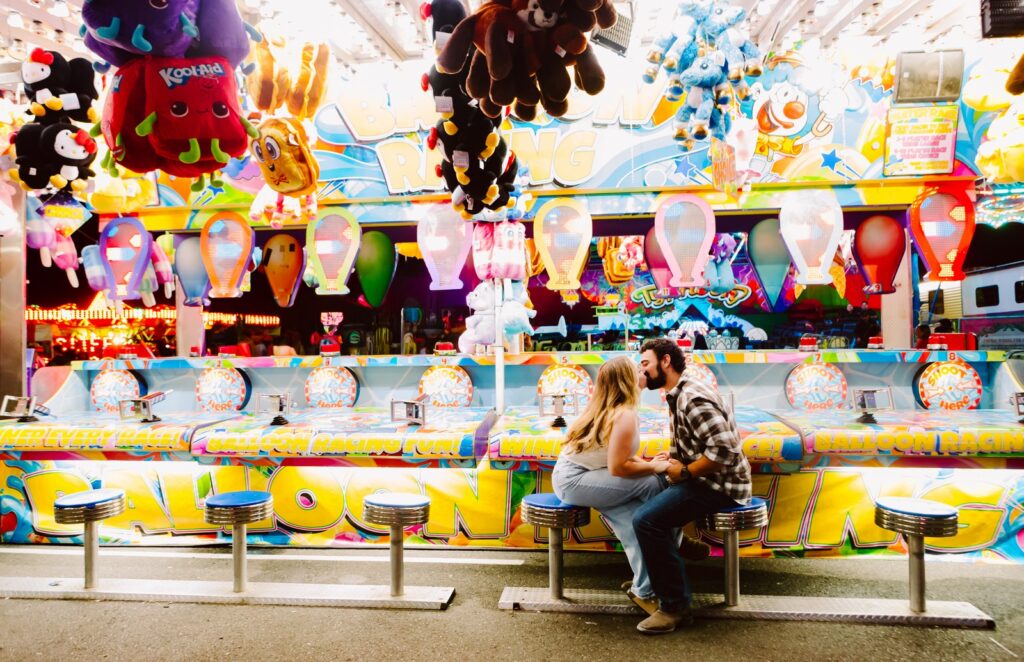 Couple kissing at a colorful carnival balloon racing game booth surrounded by stuffed animal prizes